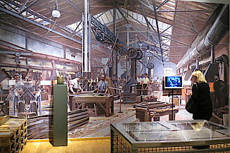 Blick in die Ausstellung: Eine Besucherin steht vor einer großen Reproduktion, die eine Werkshalle zeigt (Foto: KdFS)
