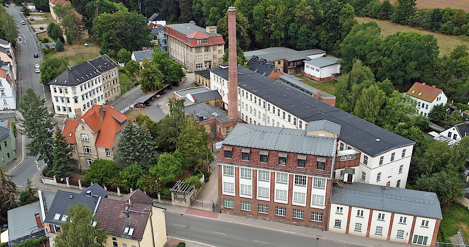 Das Foto zeigt das gesamte Gelände der Tuchfabrik Gebr. Pfau. Es ist ein Luftbild, welches mit einer Drohne aufgenommen wurde. Im Mittelpunkt ist der Schornstein zu sehen, umgeben von den verschiedenen Gebäuden der Fabrik. 