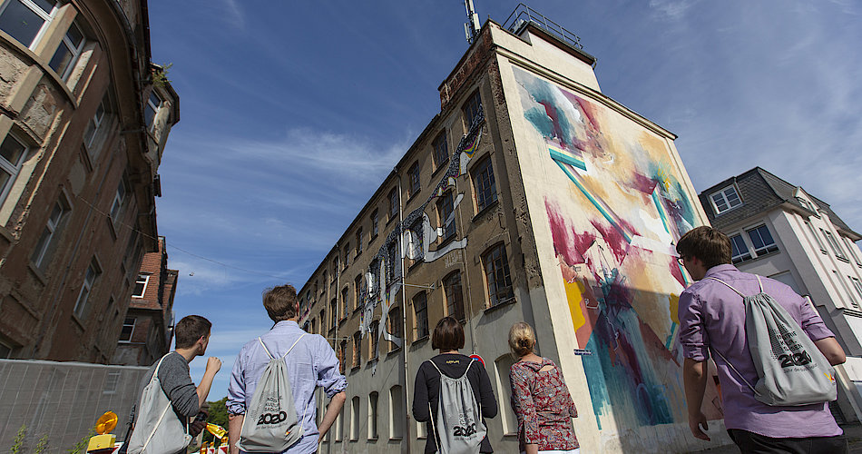 Fünf Teilnehmer einer Rallye stehen vor einem alten Fabrikgebäude. An dessen Fassade befinden sich große Bilder. Es handelt sich um Überreste der IBUG, die 2014 in Crimmitschau stattfand.